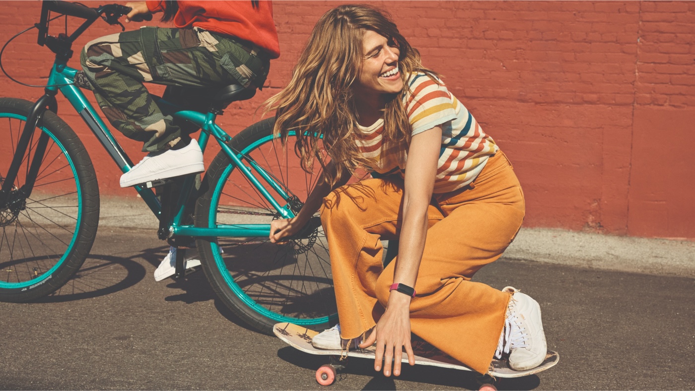 Smiling woman in orange flares and striped t-shirt, crouched on a skateboard.