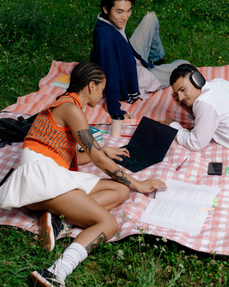 Three relaxed people lounging on a pink gingham blanket, with a laptop, notes and headphones.